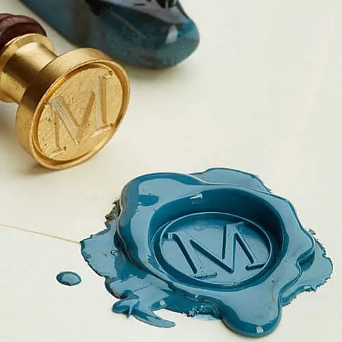 Sinete dourado com monograma “M” e lacre azul derretido com impressão personalizada.